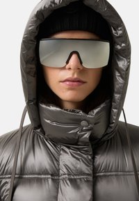 Srebrna kurtka puchowa z wysokim kołnierzem i zapięciem na guziki, w połączeniu z dużymi, lustrzanymi okularami przeciwsłonecznymi i czapką typu beanie z czarnej wełny.