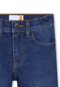 Vaqueros de mezclilla en azul oscuro con costuras doradas en contraste, que cuentan con un cierre frontal de botón, cinco bolsillos y una tela texturizada.