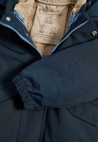 Chaqueta de color azul marino, resistente al agua, con forro de forro polar texturizado. Cuenta con un cierre de cremallera con acentos azules y dos bolsillos frontales. Etiqueta visible.