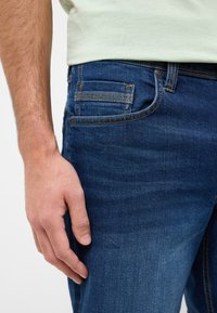 Niebieskie dżinsy o slim fit, z pięcioma kieszeniami, żółtym stębnowaniem i gładką fakturą. Ręka spoczywa na udzie.