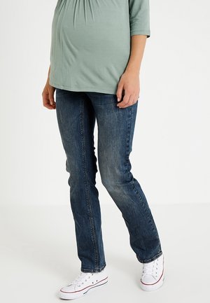 Gravid person iført lysegrøn top, blå jeans og hvide sneakers står mod en ensfarvet lys baggrund.