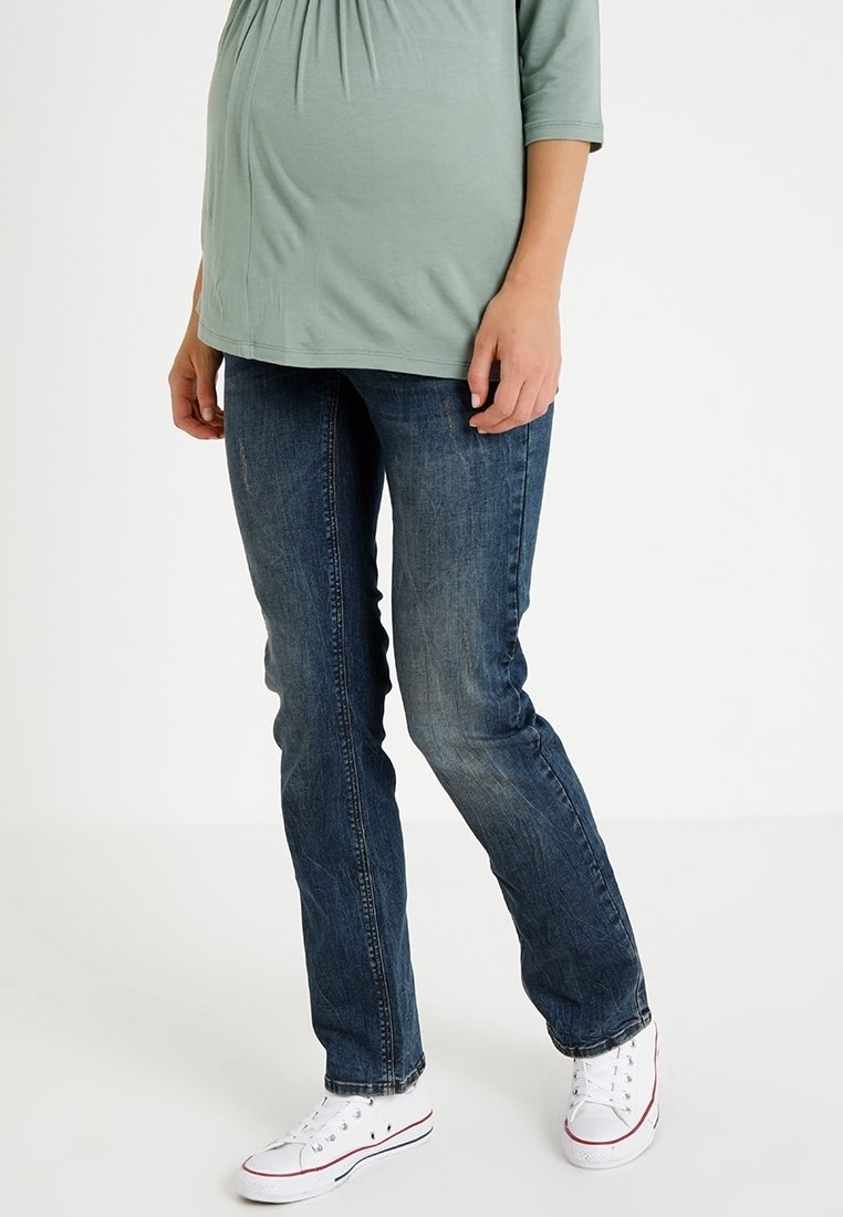 Personne enceinte portant un haut vert clair, un jean bleu et des baskets blanches, debout devant un fond clair uni.