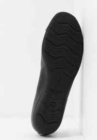 Semelle d'une chaussure noire avec des rainures ondulées, marquée "Gabor" et "Hovercraft", posée verticalement contre un mur blanc d'angle.