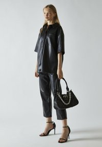 Camisa de piel sintética negra de gran tamaño con mangas cortas, combinada con pantalones ajustados a juego y un bolso negro con detalle de cadena.