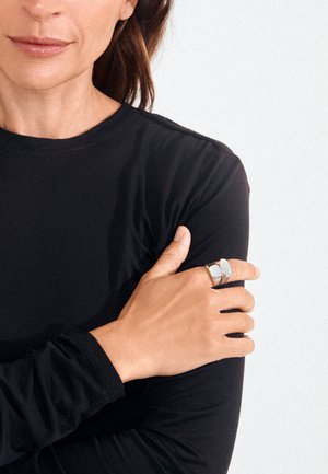 Kobieta w czarnej koszulce z długim rękawem, krzyżująca ramiona, nosząca dwa srebrne pierścionki na lewej dłoni na jednolitym jasnym tle.