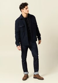 Giacca in fleece blu navy con due tasche sul petto, indossata sopra una camicia nera. Abbinata a jeans in denim scuro e scarpe in suede marroni.