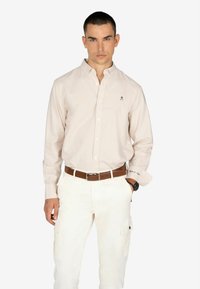 Camisa de cuadros rosa claro con mangas largas, cuello abotonado y detalle de logo. Combinada con pantalones cargo blancos y un cinturón marrón.