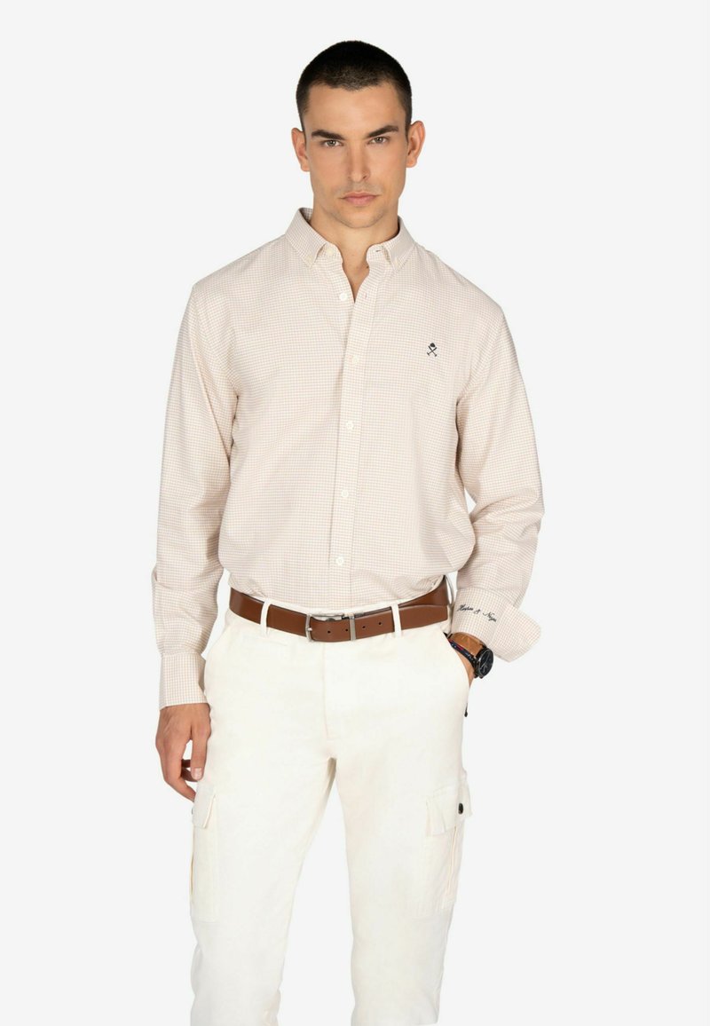 Camisa de cuadros rosa claro con mangas largas, cuello abotonado y detalle de logo. Combinada con pantalones cargo blancos y un cinturón marrón.