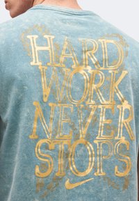 Πίσω μέρος ενός ξεθωριασμένου μπλε πουκάμισου με μεγάλα χρυσά γράμματα που γράφουν «HARD WORK NEVER STOPS» και μικρό λογότυπο της Nike από κάτω.