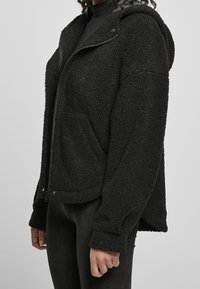 Veste en polaire noire zippée avec capuche, présentant une texture douce, des poches kangourou et des accents à boutons-pression sur le col et les poignets.