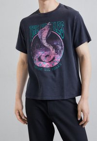 Camiseta de algodón negra con un gráfico de serpiente en tonos rosa y turquesa, con el texto "THE KOOPLES" arriba y "ABOVE ALL RULES" abajo.
