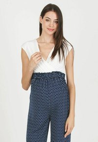 Combinaison à pois navy avec un haut blanc, présentant un design enveloppant et une taille élastique. Manches courtes à emmanchures en forme de cap et texture lisse partout.