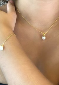 Goldene Halskette und Armband mit einer einzelnen hängenden Perle, vor Haut gesetzt, die glatte Texturen und ein einfaches Design betont.