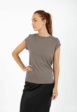 Vrouw met bruin haar in een paardenstaart draagt een taupekleurige top met capuchons en een zwarte rok, staand met één hand op haar heup, kijkend naar links tegen een witte achtergrond.