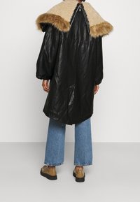 Cappotto di pelle nera con collare foderato in pelliccia beige, con vita stretta e maniche ampie, abbinato a jeans blu e scarpe marroni.