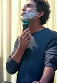 Rasoio elettrico con un design blu e nero nella mano dell'utente, mentre applica crema da barba su un viso liscio, indossando una camicia a maniche lunghe grigia.