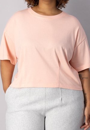 T-shirt en coton rose clair, à manches courtes et cropped, avec un col rond et une coupe décontractée, associé à un pantalon de survêtement gris.