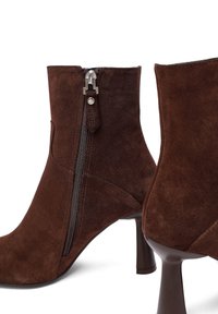 Stivali alla caviglia in suede marrone con chiusura a zip, design aderente, punta affusolata e tacco sottile. Superficie texturizzata con dettagli lisci.
