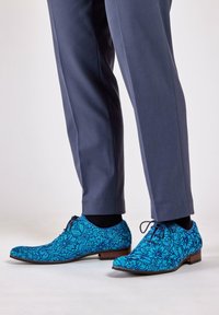 Blue floral-patterned dress shoes with a smooth texture, lace-up design, and a low heel. Paired with gray tailored trousers.