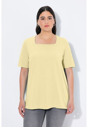 Frau mit dunklem Haar in einem blassgelben Kurzarm-Top und grauen Jeans steht vor einem weißen Hintergrund und blickt frontal in die Kamera.