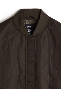 Veste Vans matelassée marron foncé avec col côtelé et fermeture frontale cachée, montrant le haut de la poitrine et l'étiquette à l'intérieur du col.