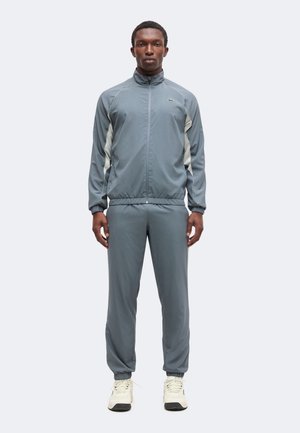 Homme debout face à l'avant, portant un survêtement gris zippé avec des panneaux latéraux blancs et des baskets blanches sur un fond uni.