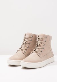 Beige suede ankle boots with round toe, padded collar, silver eyelets, and beige laces. White rubber sole with textured tread pattern.