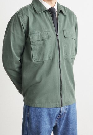 Homme portant une veste verte zippée avec de grandes poches devant, une chemise blanche, une cravate noire et un jean bleu sur un fond uni.