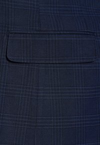 Granatowy materiał o subtelnym, teksturowanym splocie. Posiada prostokątną kieszeń na klapie oraz słabym wzorze w kratę z niebieskimi akcentami.