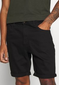 Černé džíny šortky s ohrnutými manžetami, obsahující boční kapsy a zapínání na knoflík. Nosené s tmavozelenou košilí. Texturovaná tkanina.