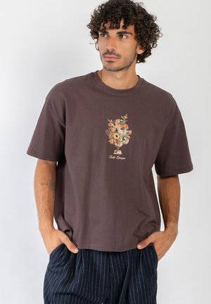 Mężczyzna z kręconymi włosami, ubrany w brązowy t-shirt z nadrukiem kwiatowego wazonu i tekstem "Belle Epoque,", ręce w kieszeniach, patrzący w bok.