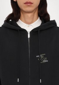 Crna dukserica s patentnim zatvaračem i vezicama, s srebrnim patentnim zatvaračem i zelenim grafičkim logotipom na prsima, preko bijele košulje s kragnom. Glatka tekstura.