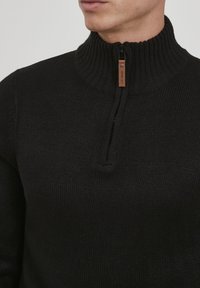 Pull en maille noire avec un col côtelé haut, doté d'une fermeture zippée à moitié et d'une étiquette en cuir sur le zip.