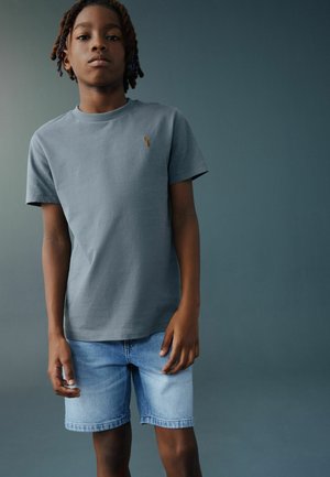 Junge mit geflochtenem Haar trägt graues T-Shirt und hellblaue Jeansshorts, steht vor einfarbigem dunkelgrauem Hintergrund.