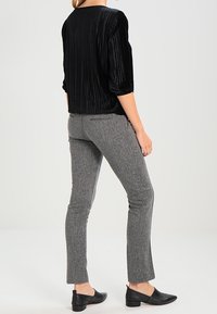 Schwarze plissierte Bluse mit Dreiviertelärmeln, kombiniert mit grauen, strukturierten, figurbetonten Hosen und schwarzen flachen Schuhen. Rückansicht, neutraler Hintergrund.