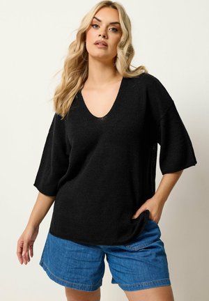 Blonde Frau trägt einen lockeren schwarzen Pullover mit V-Ausschnitt und blaue Jeansshorts, steht mit einer Hand in der Tasche vor einfarbigem Hintergrund.