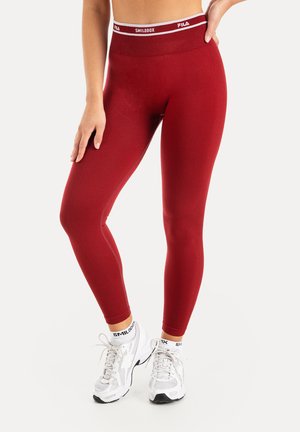 Kvinde iført røde højtaljede leggings med hvid elastisk taljebånd og hvide sportssko stående foran en ensfarvet baggrund.