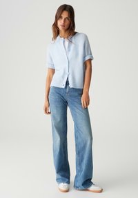 Lichtblauwe gebreide cardigan met opgerolde mouwen, knoopsluiting en een zachte textuur, gecombineerd met loszittende lichtblauwe jeans en witte sneakers.