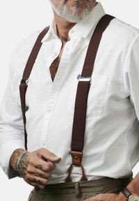 Rudi elastingi petnešos su metaliniais spaustukais ir rudais odiniais akcentais, uždėti ant baltos marškinėlių, su ryškiais tatuiruotėmis matomomis.