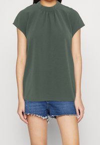 Top verde oliva a maniche corte con colletto alto e dettaglio arricciato sul davanti, abbinato a pantaloni corti di jeans strappati.