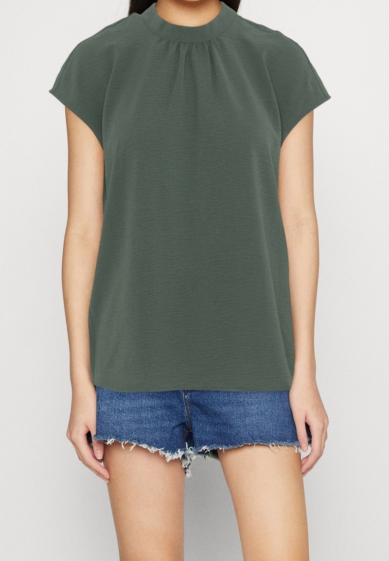 Top verde oliva a maniche corte con colletto alto e dettaglio arricciato sul davanti, abbinato a pantaloni corti di jeans strappati.