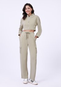 Pasukama zip-up striukė ir plačios cargo kelnės šviesiai khaki spalvos. Lygus audinys, reguliuojamas juosmuo, dvi cargo kišenės ant kelnių.