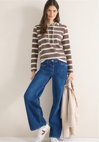 Hoodie rayé marron et blanc, jean large bleu, et baskets blanches avec des accents rouges ; mannequin tenant une veste beige texturée.