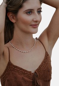 Naszyjnik składa się z naprzemiennie ułożonych pereł i małych złotych koralików, noszony na brązowym, teksturowanym ubraniu z koronkowymi zdobieniami.