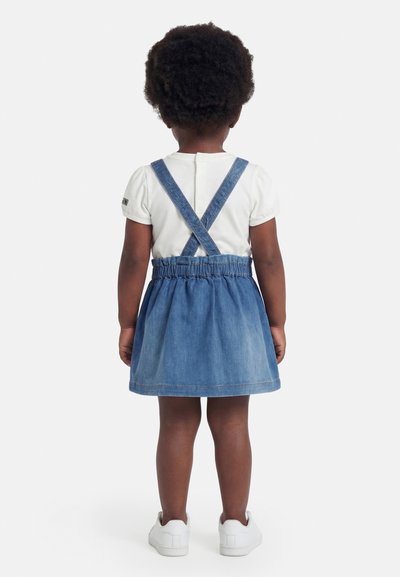 Tout-petit aux cheveux courts et bouclés, portant une chemise blanche à manches courtes et une jupe en jean bleu avec des bretelles croisées, debout de dos.