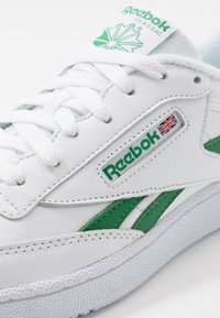 Beli usnjeni športni copat z zelenimi akcenti, ki vključuje teksturiran gumijast podplat, vezalni sistem in Reebok blagovno znamko na strani in jeziku.