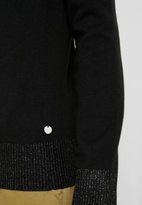 Zwarte trui met een getextureerde ribbelzoom in een contrasterend donkergrijs met zilveren draden, vergezeld van een klein metalen merkteken.