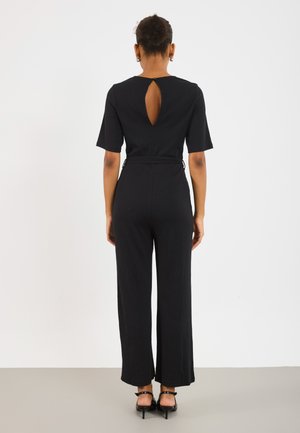 Black jumpsuit with short sleeves, ribbed texture, keyhole detail at the back, and wide legs. Worn with strappy black heels.