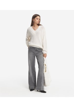 Grå utsvängda jeans i kombination med en krämfärgad, löst stickad V-ringad tröja. Outfiten inkluderar en ljus, vadderad jacka som hålls i vänster arm.