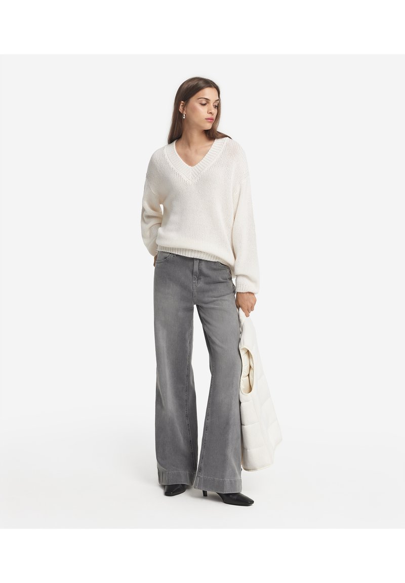 Grå, udvidede jeans parret med en cremet, løst strikket v-hals sweater. Outfit'et inkluderer en let, quiltet jakke, som holdes i venstre arm.